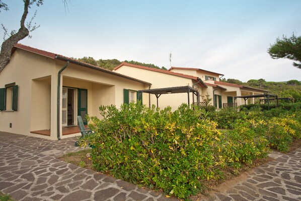 A row of beige buildings with green shutters, a stone-paved walkway, and lush greenery.