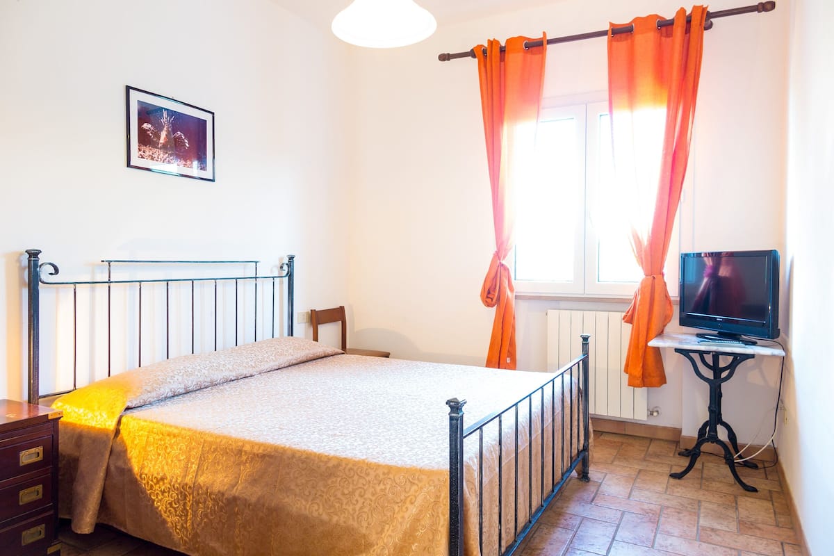 Een slaapkamer met een bed, een bureau met een computer, een raam met gordijnen en een foto aan de muur.