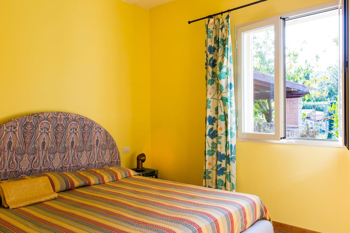 Een kamer met een bed, een raam met gordijnen en uitzicht op groen.