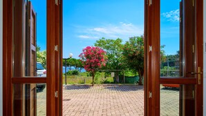 Vista a través de puertas de madera abiertas que da a un área pavimentada, un árbol en floración de color rosa, una valla y un coche aparcado al fondo.