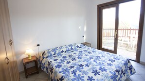 Ein Schlafzimmer mit Bett, Nachttisch mit Lampe, Fenster mit Ausblick und einer Tür.