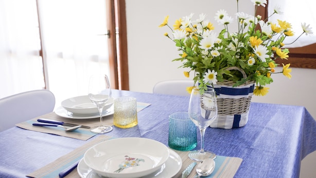 Una mesa de comedor dispuesta con platos, cubiertos y vasos, con una cesta de flores y una vela sobre la mesa.