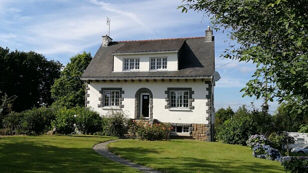 A two-story house with a stone facade, a central entrance with a door, and a small porch with a flower bed. - Chambres d'Hôtes Au Cœur De Breizh (Plouguernevel)