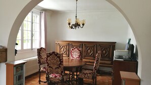 A dining area with a wooden table and chairs, a chandelier, and a view through an arched window. - Chambres d'Hôtes Au Cœur De Breizh (Plouguernevel)