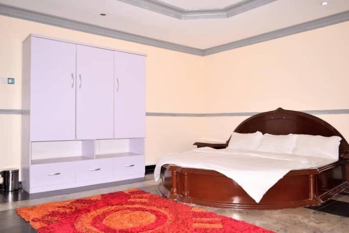 A bedroom with a large white wardrobe, a wooden bed with white bedding, and a red patterned rug.