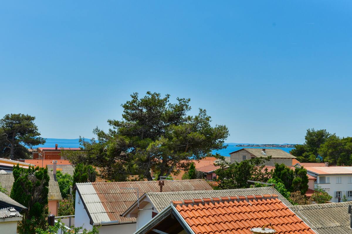A coastal residential area with terracotta-roofed houses, a prominent tree, and a clear blue sky.