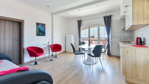 Una habitación moderna con cama, una zona de comedor con sillas, una cocina con fregadero y un amplio ventanal con cortinas.