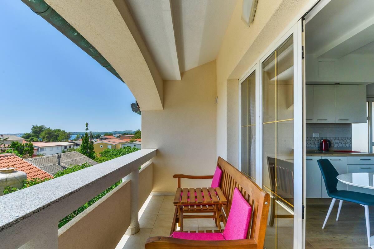 A balcony with a wooden bench and a table, a kitchen area with cabinets, and a view of the neighborhood.