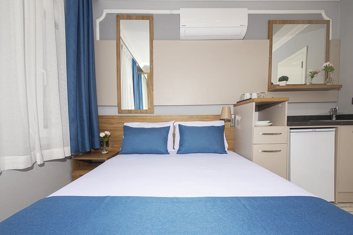 Ein Hotelzimmer mit einem Bett, einem Nachttisch mit Blumenvase, einem Spiegel, einer kleinen Kommode und einer Küchenzeile.