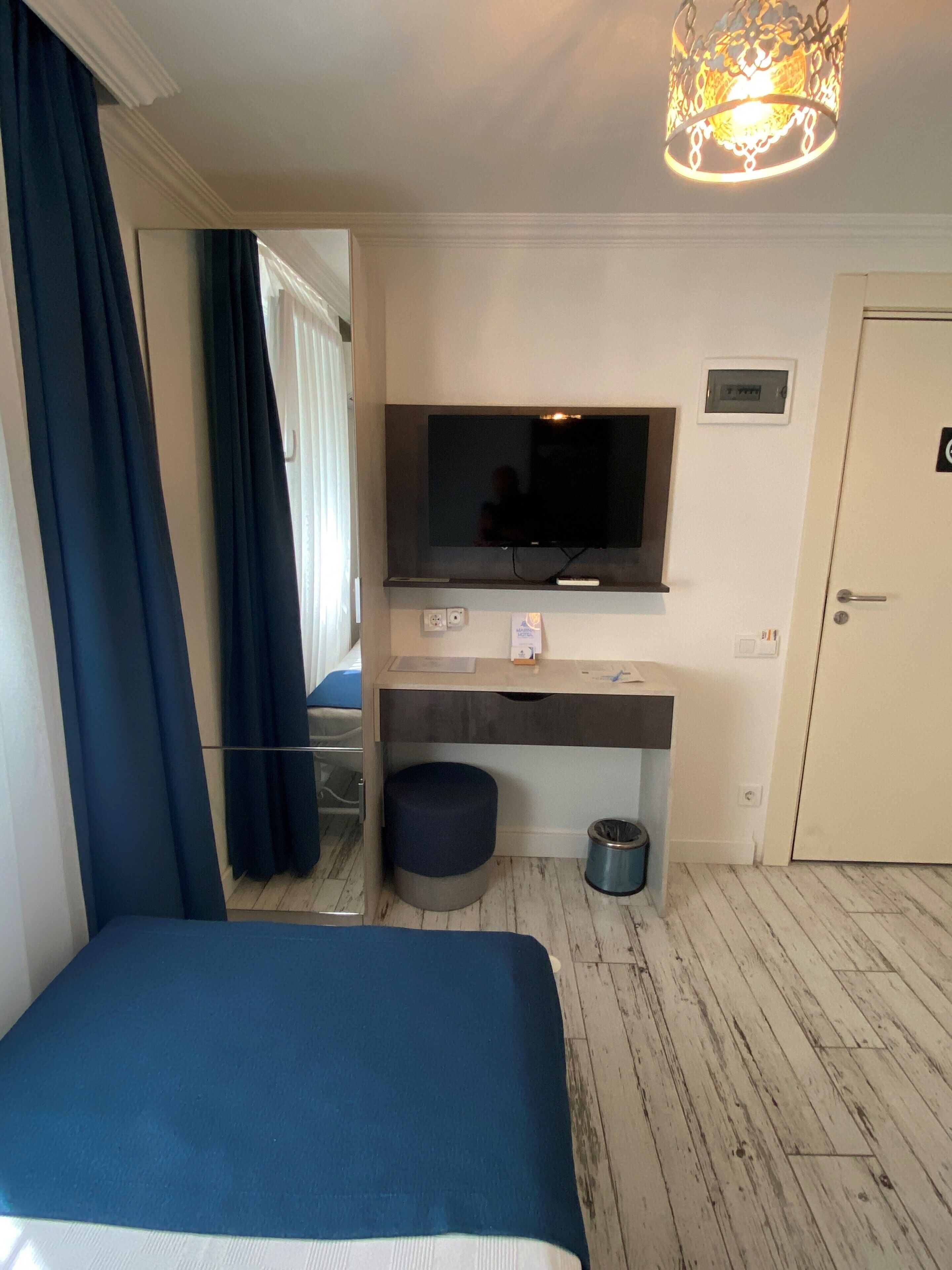 A hotel room with a bed, a TV mounted on the wall, a desk with a computer, a trash bin, and a door.