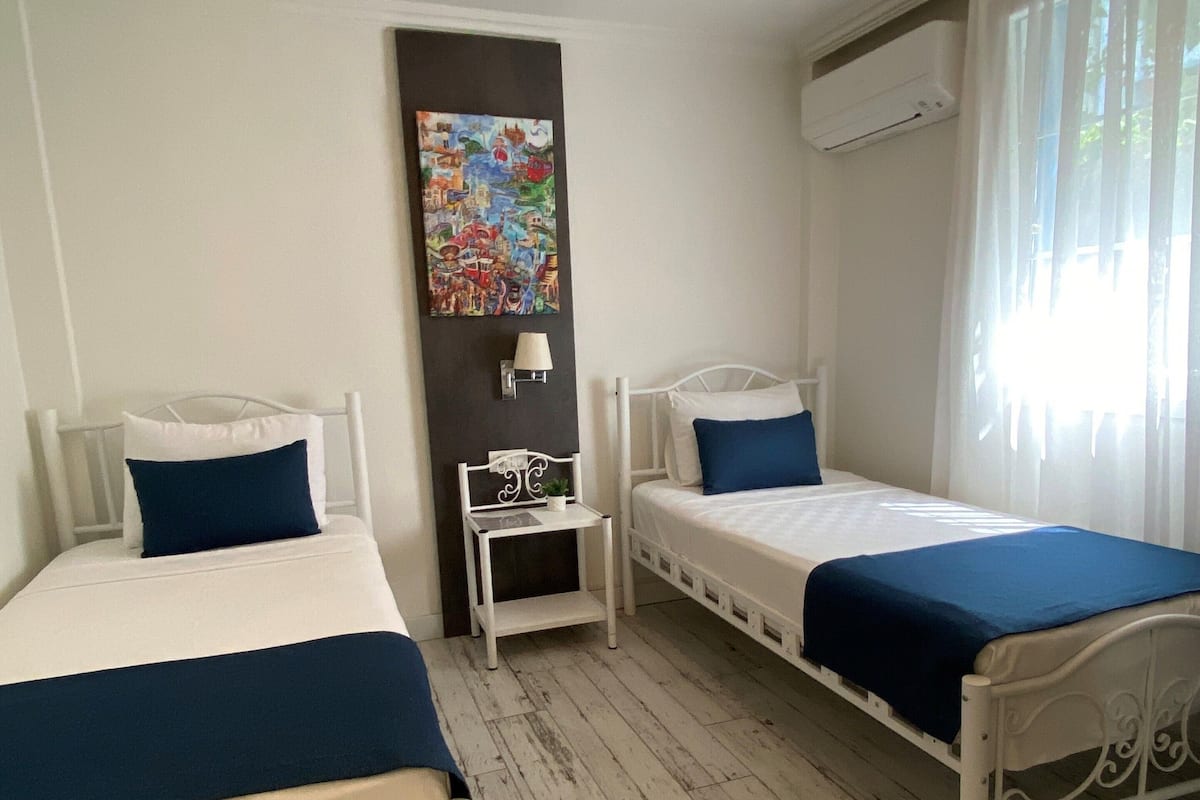 Ein Zimmer mit zwei Betten, einem kleinen Tisch, einer Wandlampe und einem Bild.