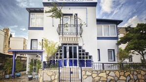 Un hotel con fachada blanca, detalles azules, un balcón y una entrada con muro de piedra.