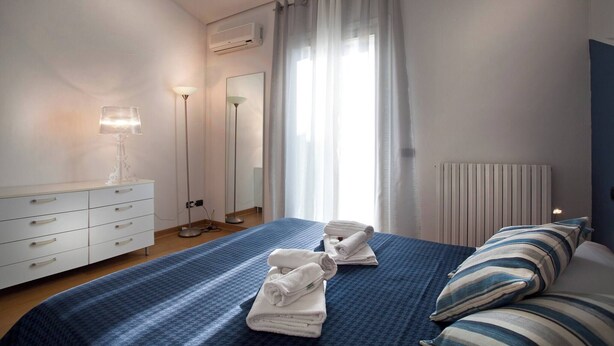 A hotel room with a bed, a bedside table with a lamp, a dresser, a radiator, and a window with curtains.