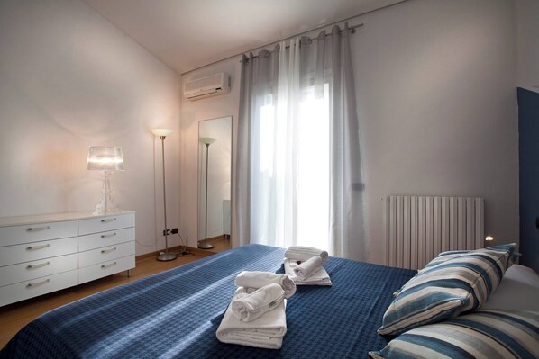 A hotel room with a bed, a bedside table with a lamp, a dresser, a radiator, and a window with curtains.