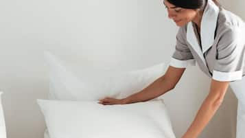 Eine Person richtet ein weißes Kopfkissen auf einem Bett in einem Zimmer mit einer schlichten, weißen Wand zurecht.