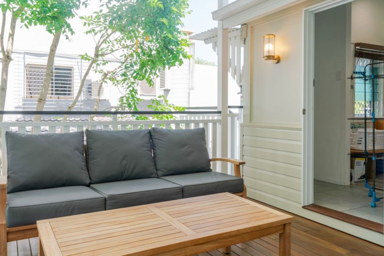 A wooden deck with a sofa and a table, a white door with a light fixture, and a view of trees and buildings.