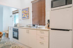 Une cuisine compacte équipée d’un four à micro-ondes, d’un four et d’un évier. On y trouve une fenêtre avec des stores, un tapis à motifs et un tableau accroché au mur.