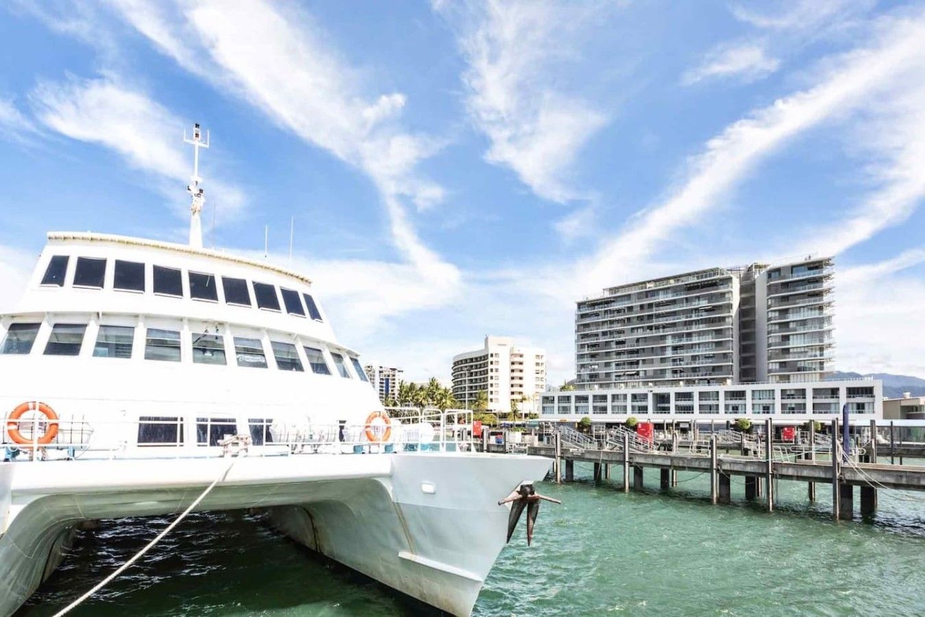 A waterfront scene with a large white boat docked near a pier, a modern building with multiple floors, and clear skies.
