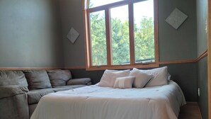 Un dormitorio con una cama grande, un sofá gris y una ventana con vistas a árboles.
