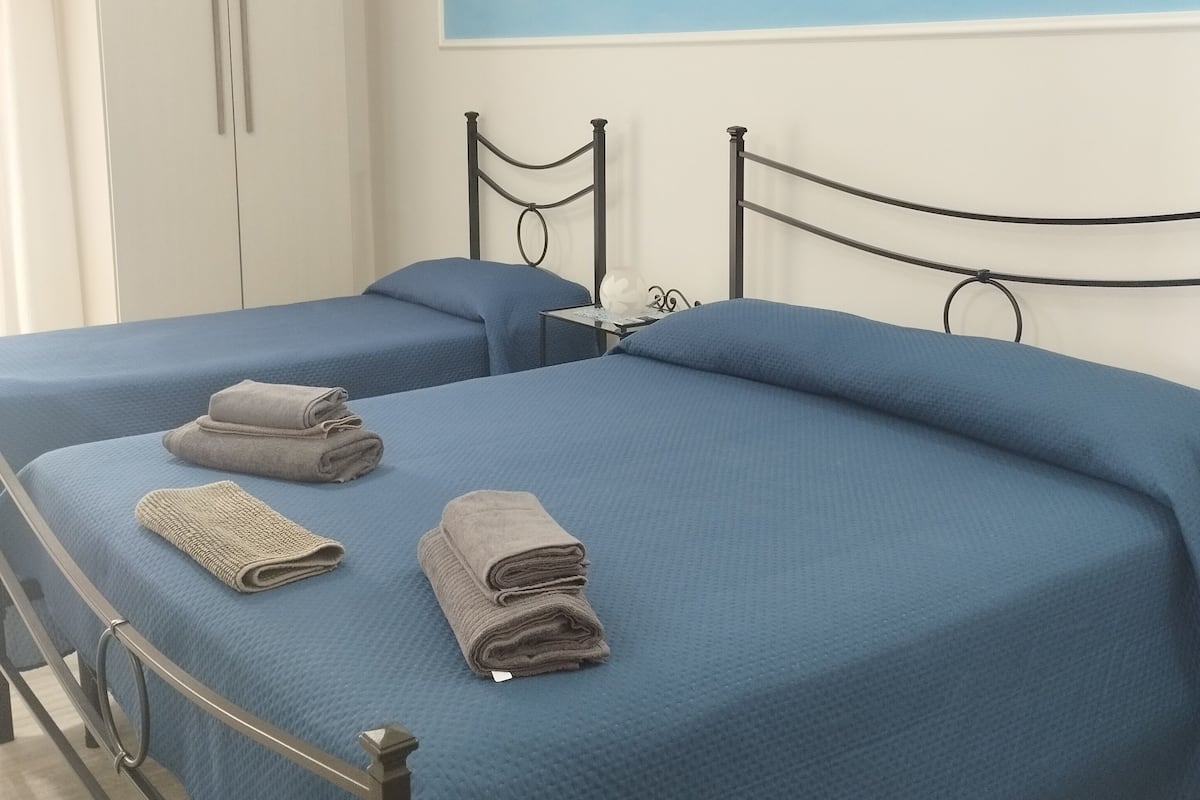 A bedroom with two beds, each with a blue bedspread and a metal headboard. There are towels on the beds and a wardrobe in the background.