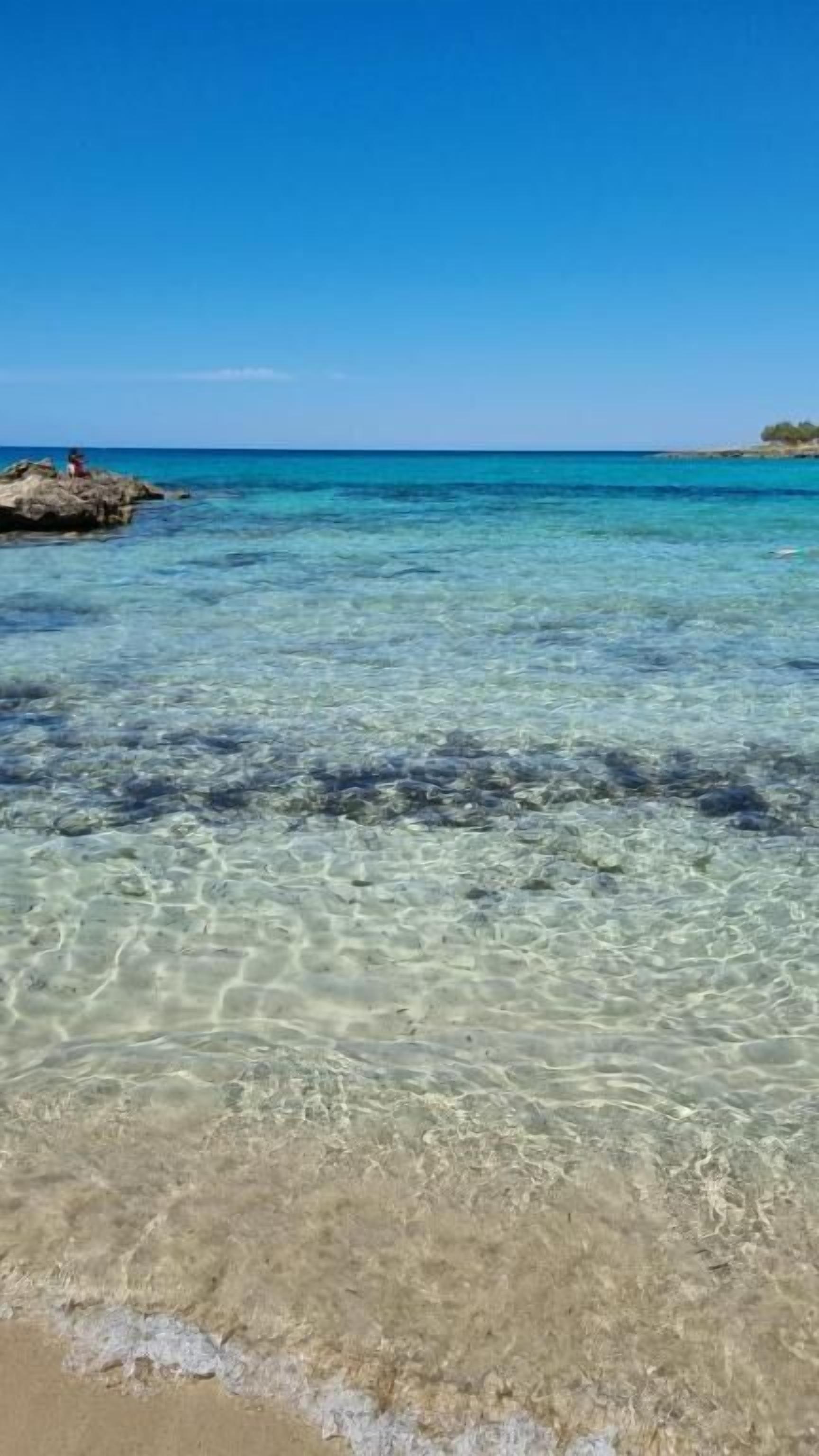 A beach with clear shallow water and a rocky outcrop.