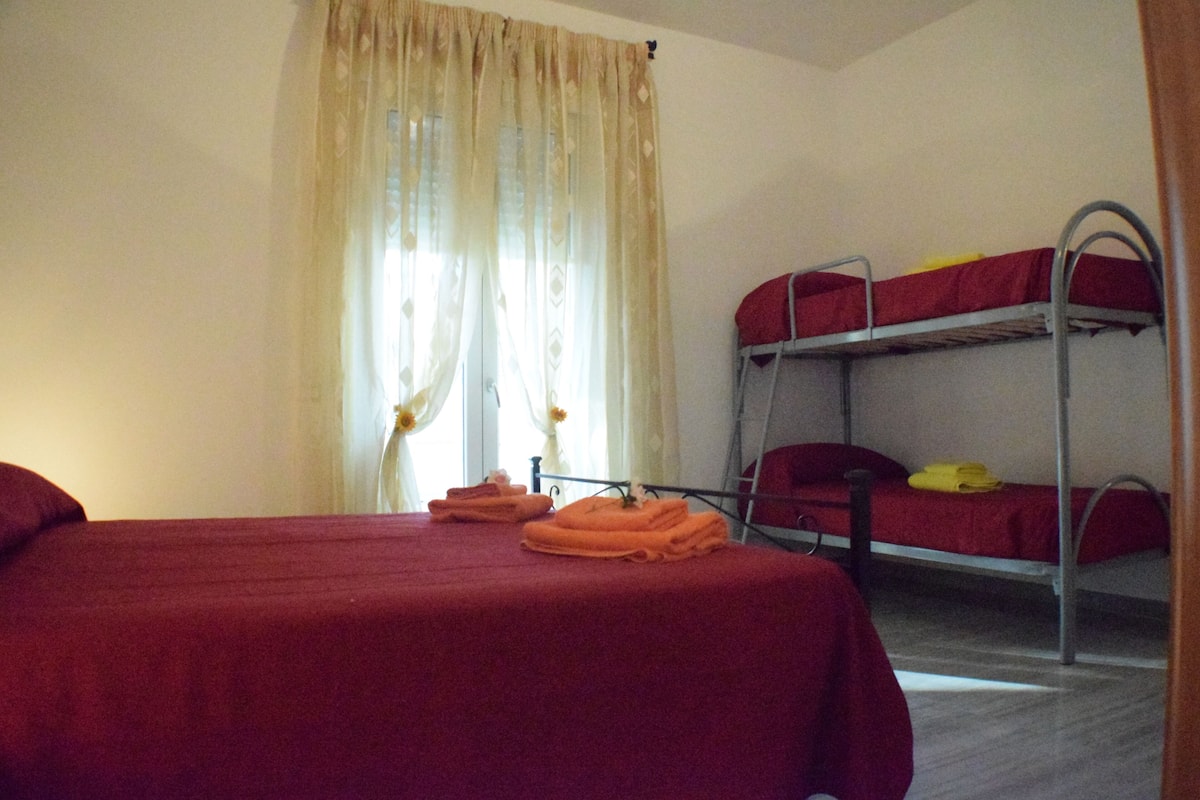 A room with two beds, one with a red bedspread and the other with a red and yellow bedspread. There are folded towels on the bed with the red bedspread.
