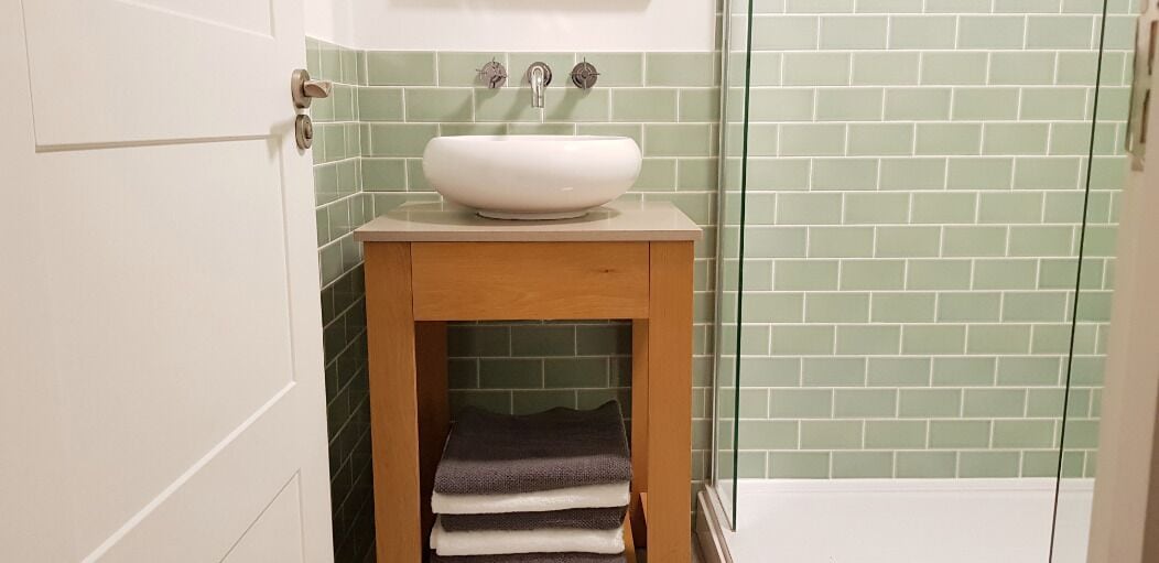 A bathroom with a white sink on a wooden vanity, green-tiled walls, and a glass shower door.
