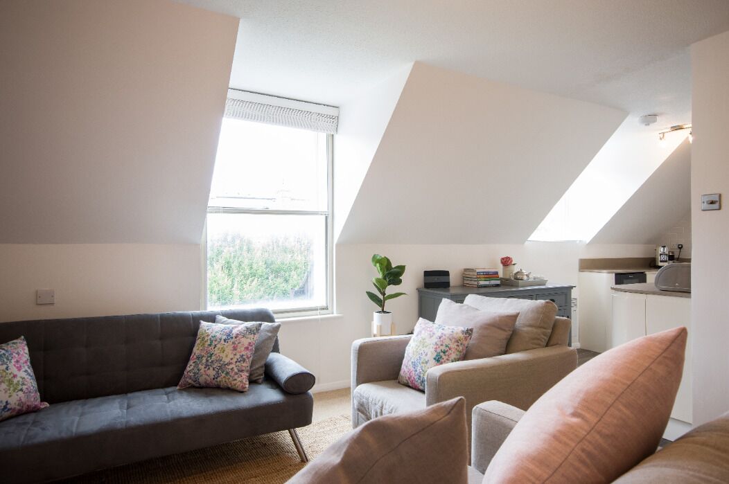 A modern living room with a gray sofa and armchairs, a small kitchen area, and a large window providing natural light.