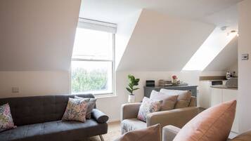 A modern living room with a gray sofa and armchairs, a small kitchen area, and a large window providing natural light.