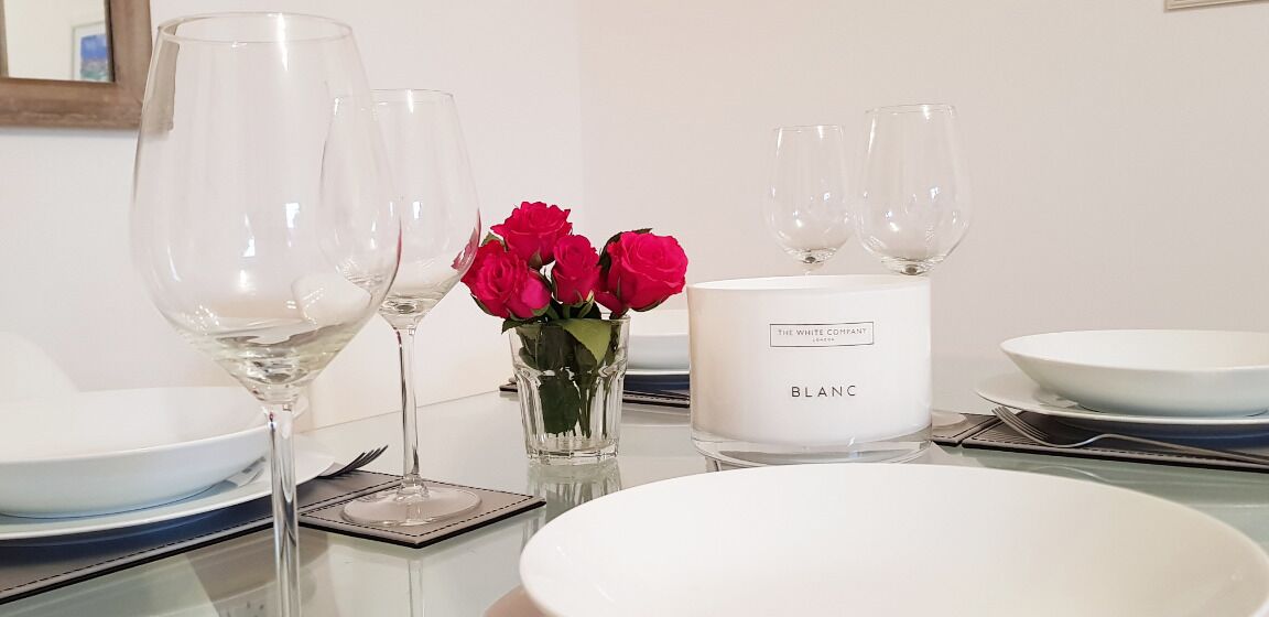 A table set with white plates, clear wine glasses, a white candle labeled "BLANC," and a vase with red roses.