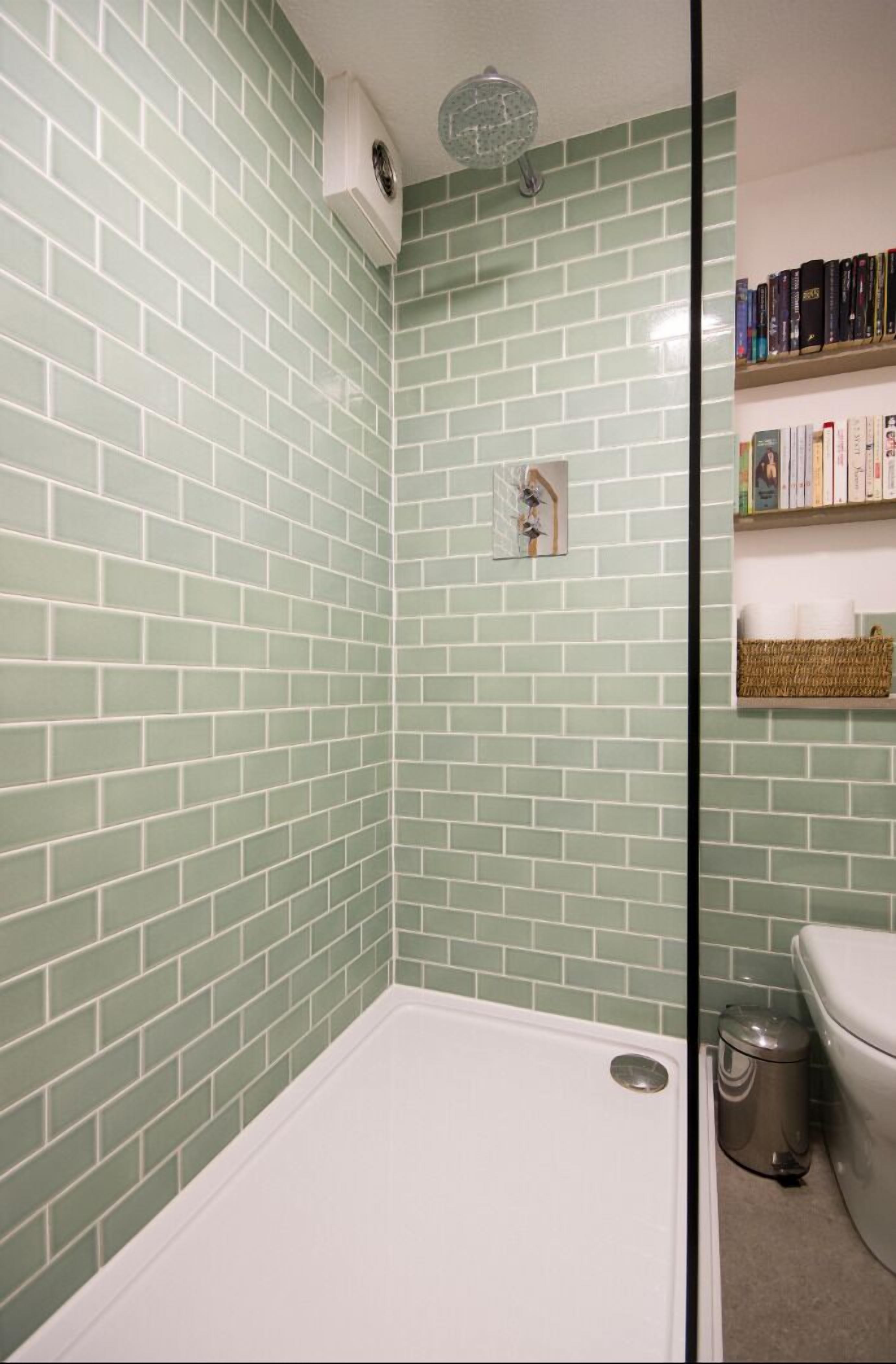 A bathroom with green-tiled walls, a white bathtub, a showerhead, a toilet, and a shelf with books and a basket.