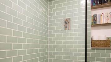 A bathroom with green-tiled walls, a white bathtub, a showerhead, a toilet, and a shelf with books and a basket.