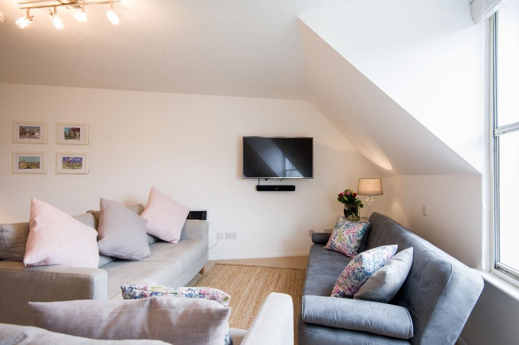 A modern living room with a grey sofa set, a flat-screen TV mounted on the wall, and a window providing natural light.