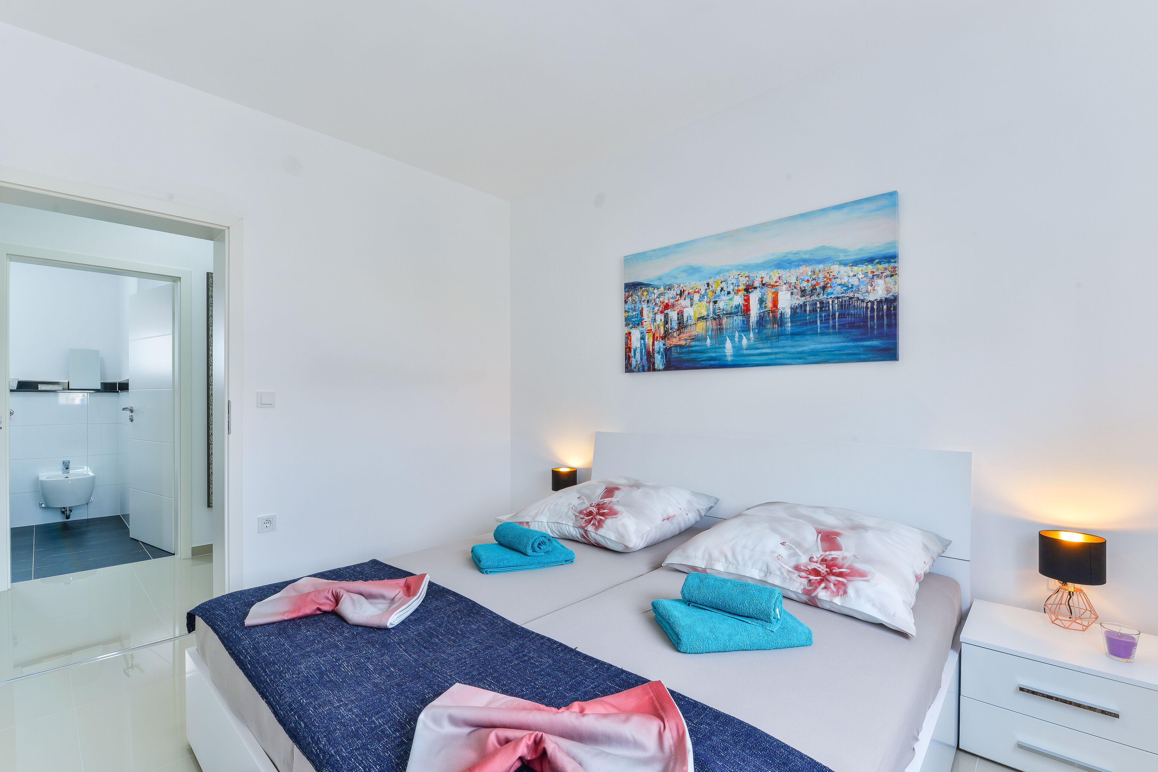 A modern hotel room with a bed, two bedside tables, a painting, and a bathroom visible through an open door.