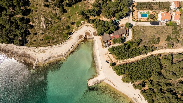 An aerial view of a coastal property with a beach, a small boat dock, and a house with a pool. - Apartments Pavlak (Vir)