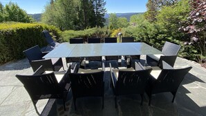 An outdoor dining area with a glass table and black wicker chairs, surrounded by lush greenery and trees. - Villa in the Black Forest with indoor pool (Falkau)