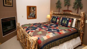 Un dormitorio con estructura de cama de madera rústica, colcha con estampados de animales, chimenea y adornos en la pared, incluyendo un espejo y árboles artificiales.