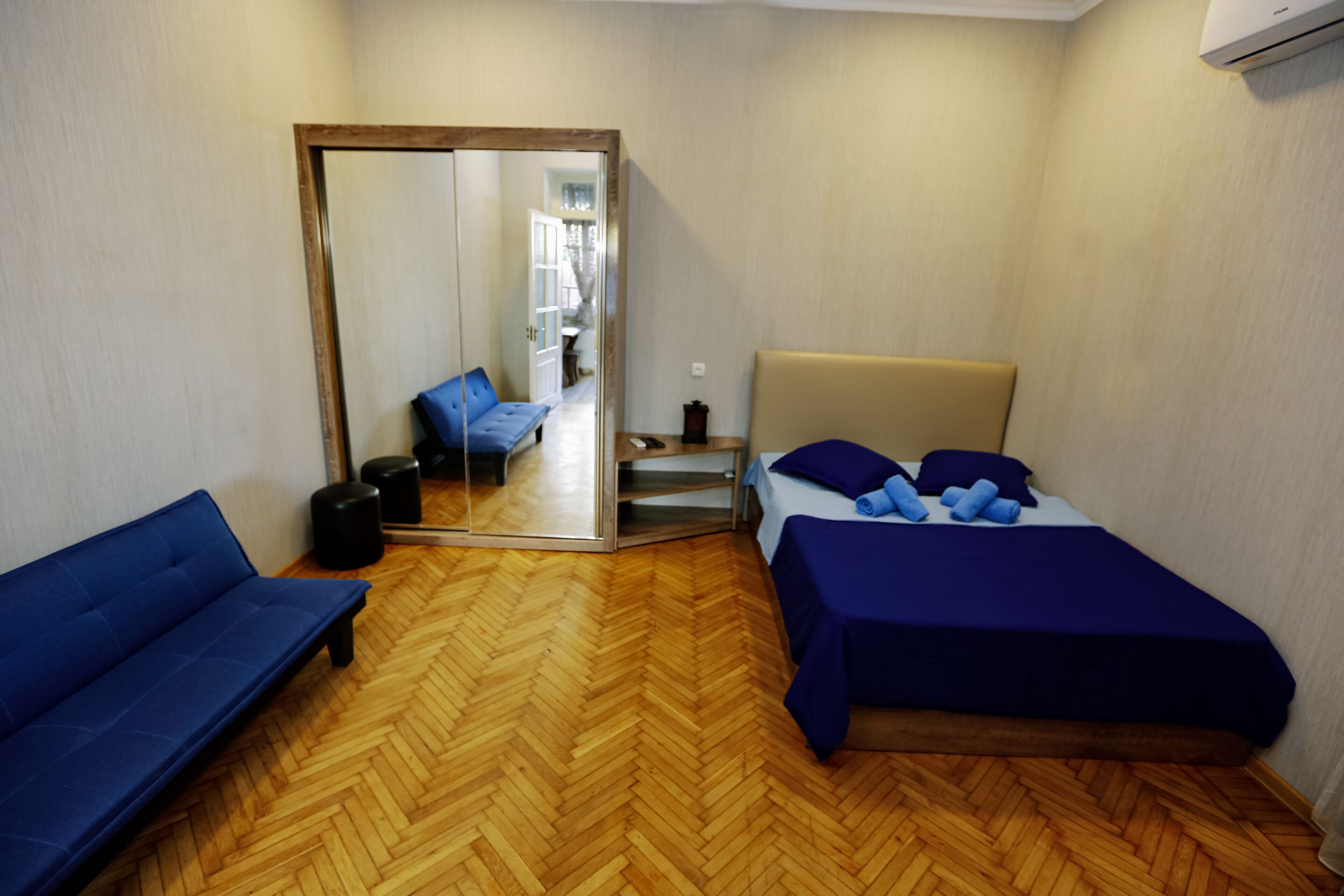 A room with a bed, a sofa, a mirror, and a wooden herringbone floor.