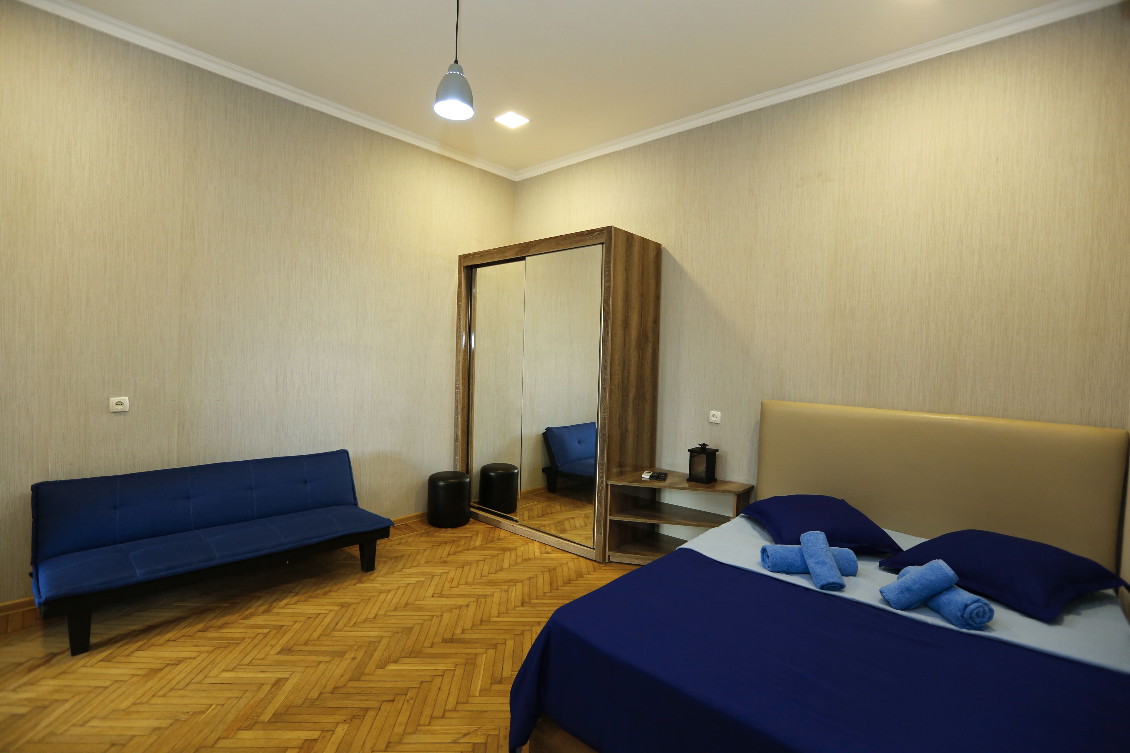 A room with a bed, a blue sofa, a mirror, and a wooden floor.