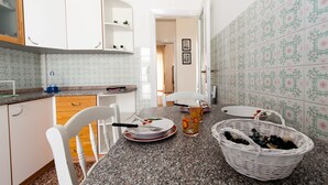 A kitchen with white cabinets, a granite countertop, and patterned tiles. There are two white chairs, a dining table with plates and glasses, and a wicker basket on the counter. - Casa Vacanza Circe (Sperlonga)