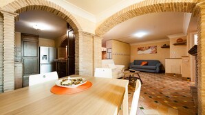 A dining area with a wooden table, chairs, and a plate of food. There is a kitchen area with a refrigerator and cabinets. The room has a tiled floor and brick walls. - Casa Vacanza Polifemo Loft (Sperlonga)