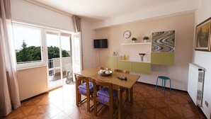 A dining area with a wooden table, chairs, a TV, a clock, and a view of greenery outside. - Casa Vacanza Santa Laura (Sperlonga)