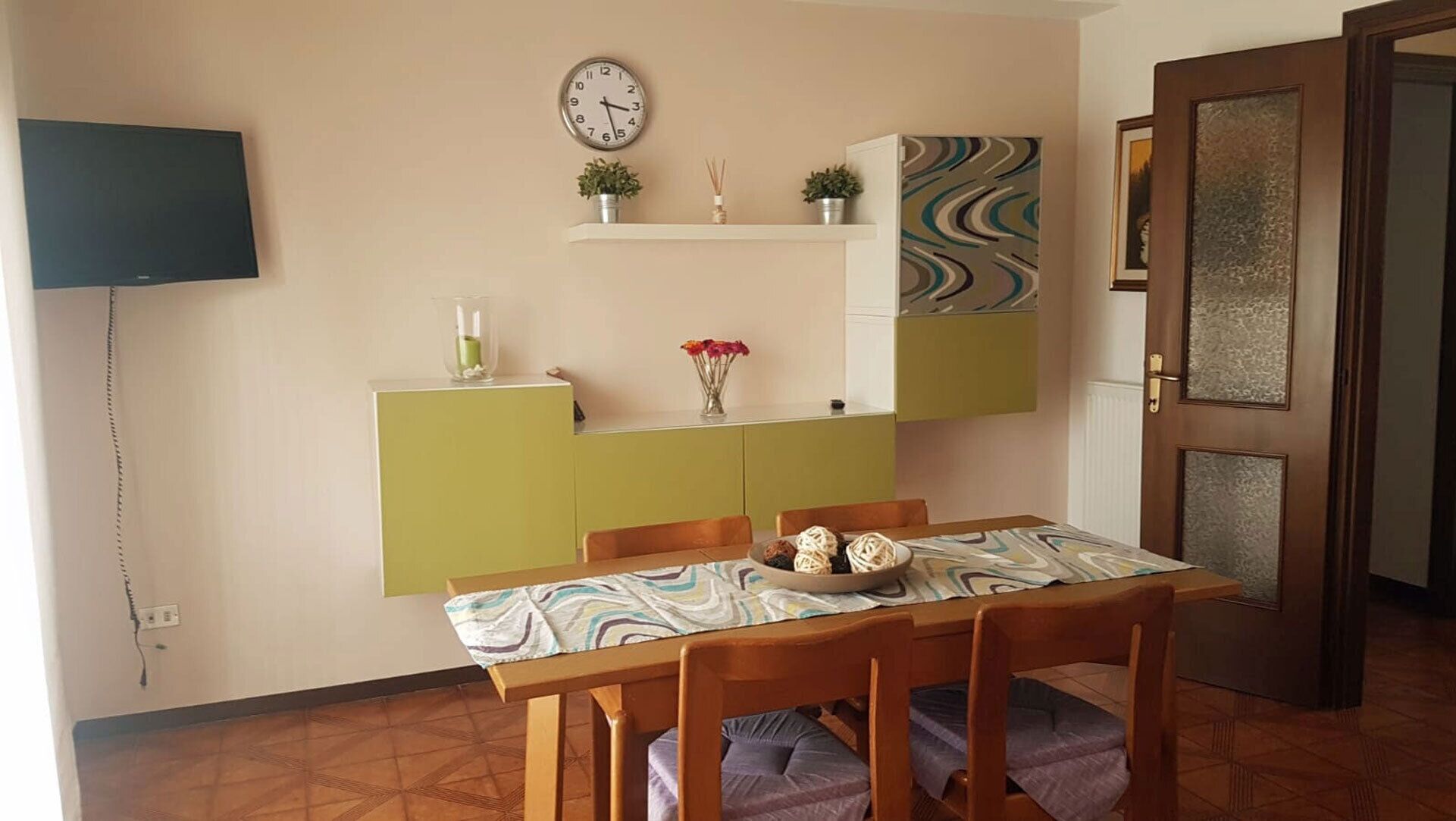 Un coin repas avec une table dressée pour deux personnes, une horloge au mur et une porte dotée d’un panneau à motifs.