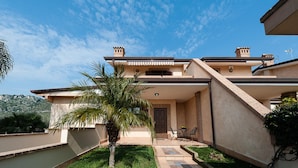 A modern two-story house with a flat roof, a small patio with a table and chairs, and a palm tree in the front yard. - Casa Vacanza Villa Polifemo (Sperlonga)