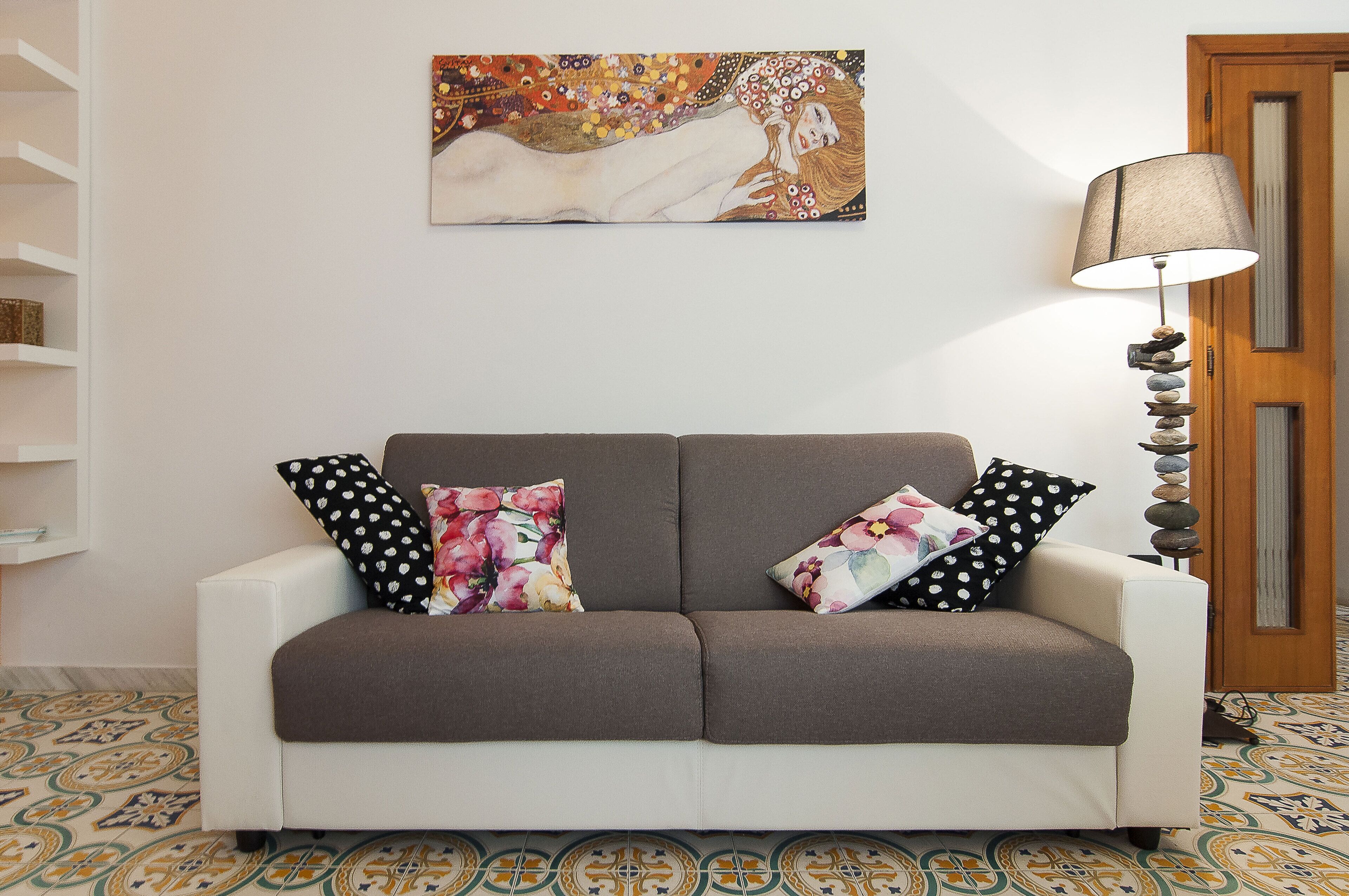A room with a sofa, pillows, a lamp, and a painting on the wall.