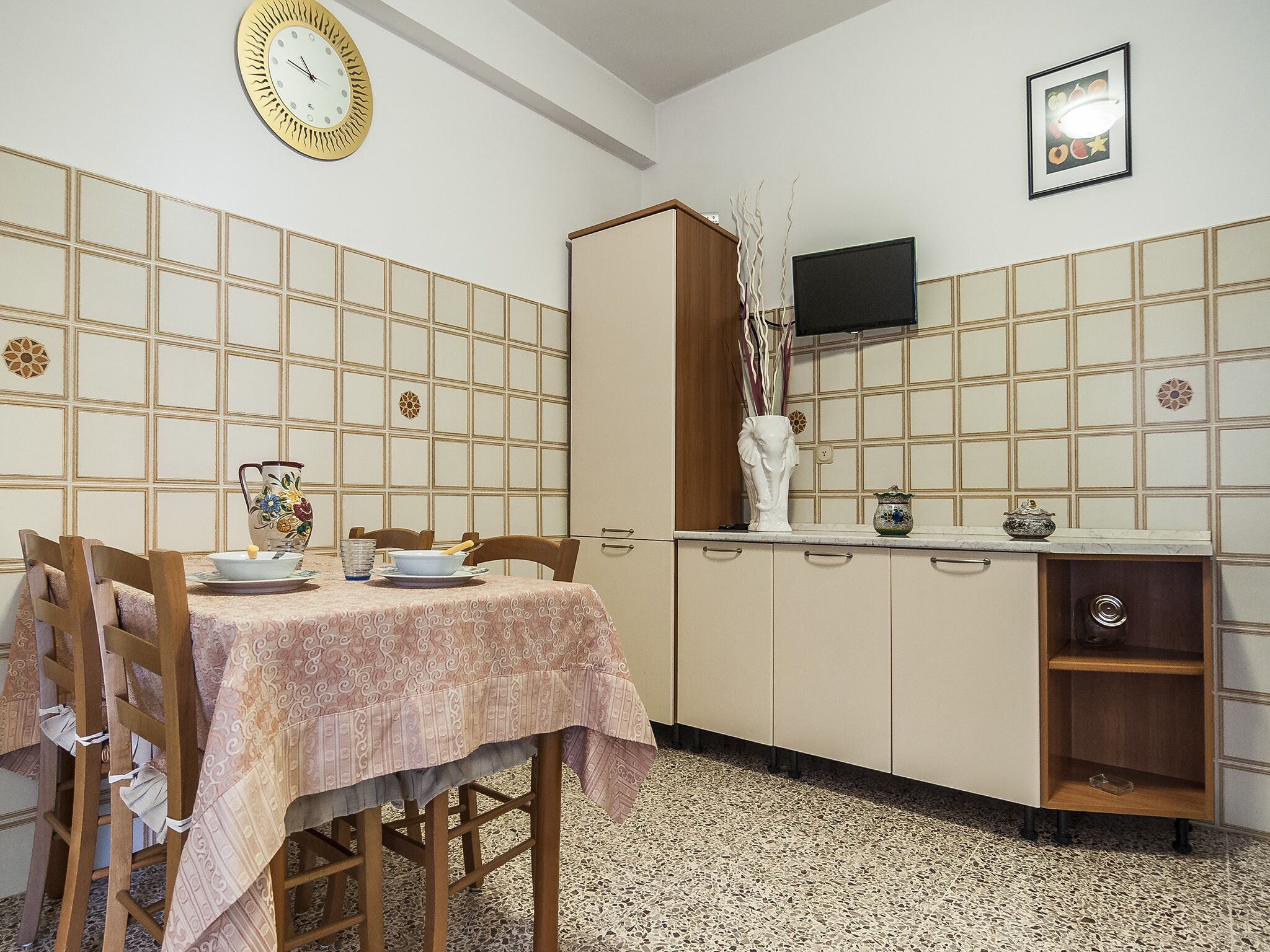 Un comedor con una mesa dispuesta para cuatro personas, un reloj en la pared y un televisor sobre un mueble.