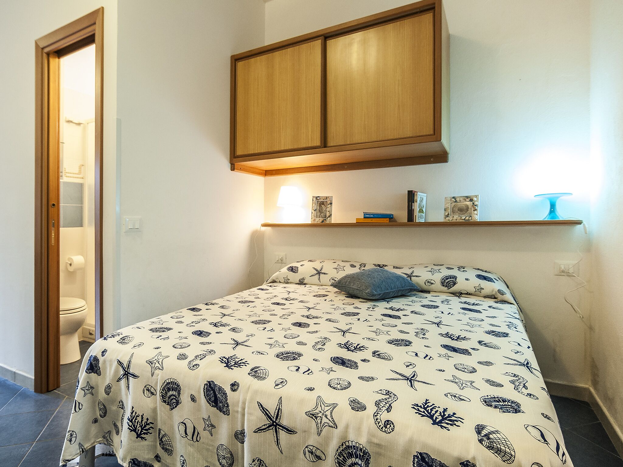 A bedroom with a bed, a wooden cabinet above the bed, a shelf with books and a lamp, and a bathroom door visible to the left.