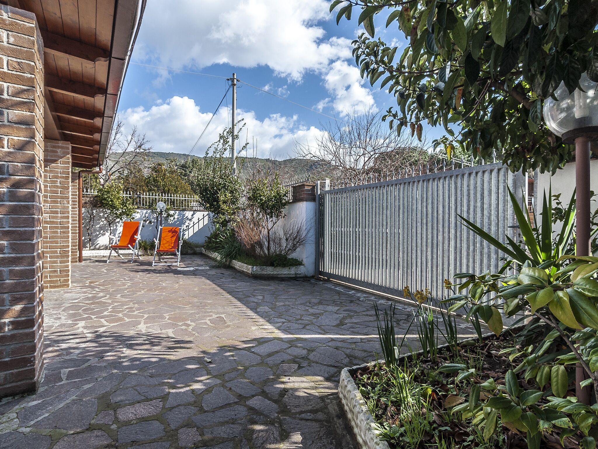 A courtyard with stone flooring, a brick wall, a metal fence, and some greenery.