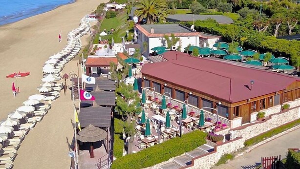 Un hotel frente al mar con mobiliario en la playa, un restaurante y vistas a la montaña.