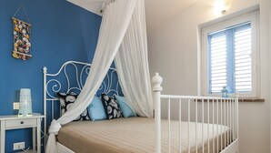 Un dormitorio con estructura de cama metálica blanca, dosel, mesita de noche con lámpara y ventana con persianas.